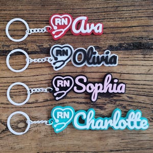 May include: Four keychains with names: Ava, Olivia, Sophia, and Charlotte. Each keychain features a white ring and chain, with a colored RN stethoscope design and the name in a matching color. The keychains are on a wooden surface.