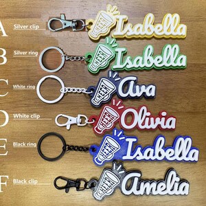 May include: Keychains with names and cheerleading megaphone designs. The keychains feature names like "Isabella", "Ava", "Olivia", and "Amelia" in various colors. Each keychain has a different clip or ring attachment.