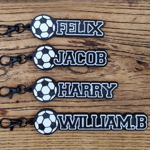 May include: Four black and white keychains featuring a soccer ball icon attached to a black lobster clasp. Each keychain displays a personalized name in white block lettering: FELIX, JACOB, HARRY, and WILLIAM.B, all set against a black background.