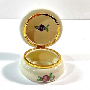 May include: A small, round, white porcelain trinket box with a gold trim. The lid is open and features a purple and white flower design. The bottom of the box has a similar flower design.