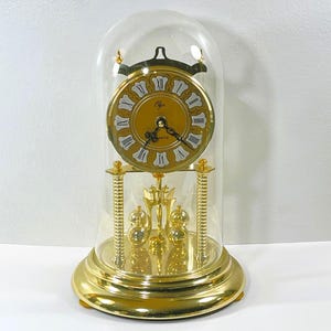 May include: A gold-toned quartz clock with a glass dome. The clock face is gold with Roman numerals and black hands. The base and decorative elements are gold. The clock is a decorative item.