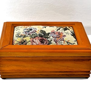 Puede incluir: Una caja de madera con un forro de tela con estampado floral. La caja tiene una tapa con bisagras y un borde decorativo alrededor de los bordes.