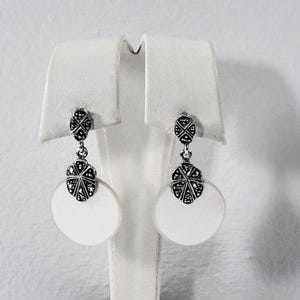 May include: A pair of drop earrings featuring a circular white disc suspended from a silver-toned metal component with a textured, dark-accented geometric pattern. The top section of each earring is a smaller, matching silver-toned piece.