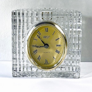 May include: A clear, square crystal quartz clock with a gold-tone face and Roman numerals. The clock face is marked "MIKASA" and "QUARTZ". The clock is a decorative item.
