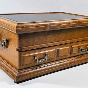 Op de afbeelding: Een houten sieradendoos met een donkere bovenkant en twee kleine lades. De doos is gemaakt van lichtbruin hout met een glanzende afwerking. Hij heeft sierlijke metalen handgrepen en staat op een verhoogde basis. De doos is rechthoekig en heeft een klassiek ontwerp.