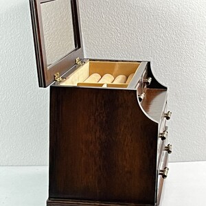 May include: A dark brown wooden jewelry box with a hinged lid and a mirror inside. The box has four drawers with gold-colored knobs. The box is sitting on a white surface.