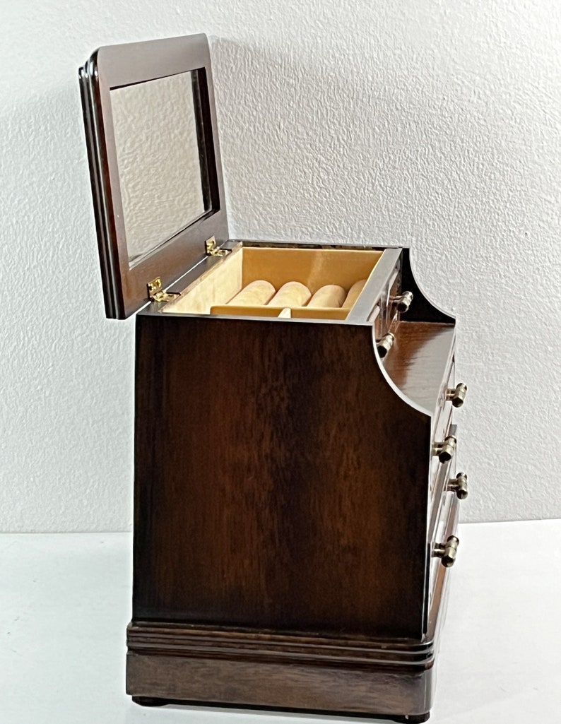 May include: A dark brown wooden jewelry box with a hinged lid and a mirror inside. The box has four drawers with gold-colored knobs. The box is sitting on a white surface.
