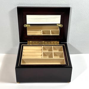 Puede incluir: Una joyero de madera marrón oscuro con tapa con bisagras y espejo incorporado. El interior presenta múltiples compartimentos y ranuras para anillos forradas con tela beige claro. Herrajes dorados acentúan la caja.