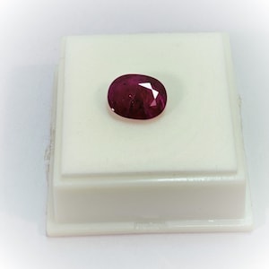 Puede incluir: Una piedra preciosa ovalada de color rojo oscuro sobre un soporte de exhibición blanco.