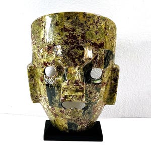 May include: A stone mask with a face-like design. The mask is primarily green with brown and black accents. It has two circular eye holes and a rectangular mouth opening. The mask is mounted on a black base.
