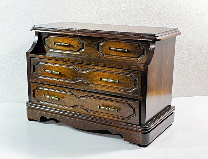 May include: A dark wood jewelry box with four drawers and brass pulls. The box has a curved front and a slightly raised top.