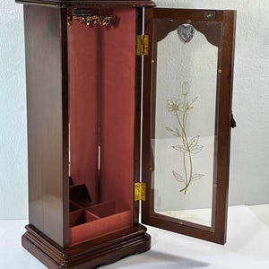 Op de afbeelding: Een hoge sieradenkast van donker hout met een open scharnierende deur voorzien van een glazen paneel met een goudkleurig bloemmotief en een klein hartje. De binnenkant is bekleed met roze stof en bevat een hakenrek en vakken aan de onderkant.