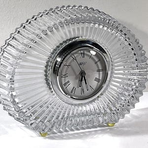 May include: A clear, glass clock with a radial design. The clock face is silver with black Roman numerals and hands. The clock has a silver bezel and sits on small, gold-colored feet.