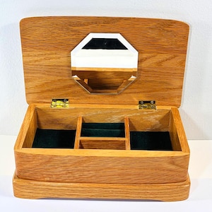Puede incluir: Un joyero rectangular de madera con acabado de veta de roble marrón claro. La tapa está abierta, revelando un espejo octogonal montado en el interior. El interior está forrado con tela verde oscuro y cuenta con tres compartimentos.