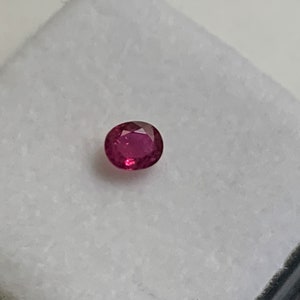 Pode incluir: Uma gema oval de cor rosa vibrante, provavelmente um rubi, exibida em uma superfície branca.