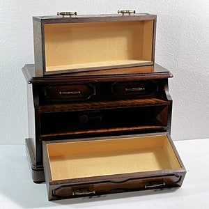 May include: A dark wood jewelry box with a hinged top and three drawers. The top drawer is open, revealing a beige interior. The bottom drawer is also open, revealing a beige interior.