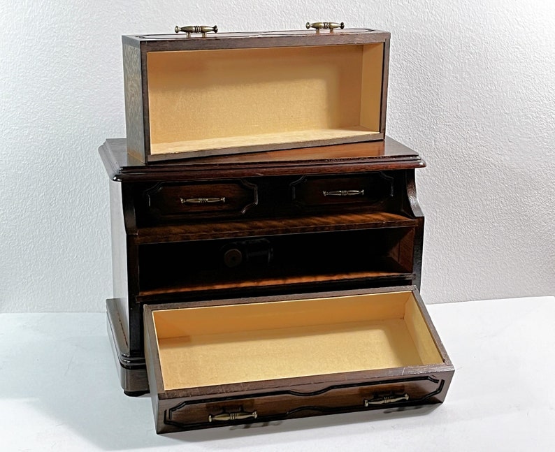 May include: A dark wood jewelry box with a hinged top and three drawers. The top drawer is open, revealing a beige interior. The bottom drawer is also open, revealing a beige interior.