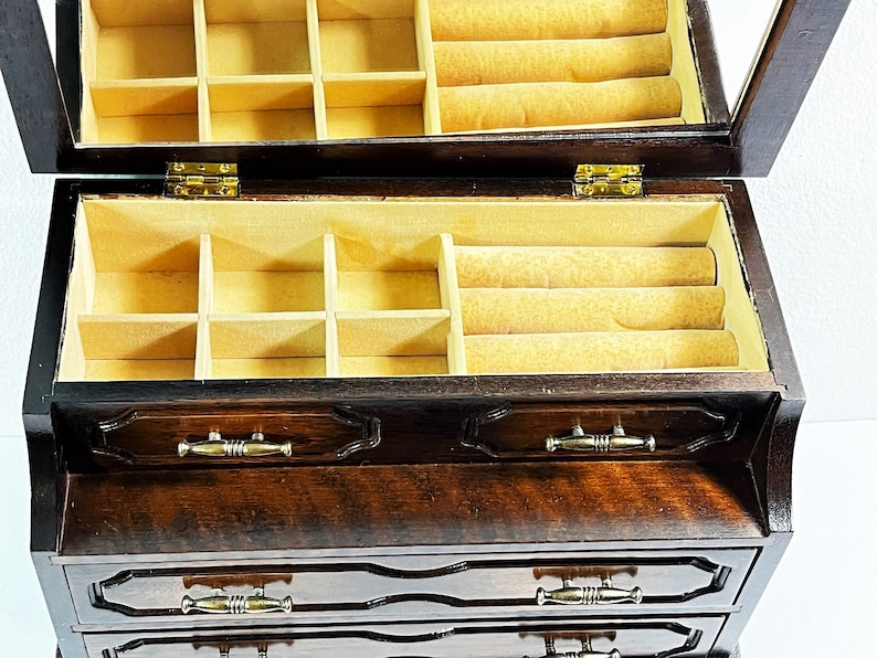 May include: A dark wood jewelry box with a hinged lid and a mirror inside. The box has four drawers and a top compartment with multiple compartments and a ring roll.