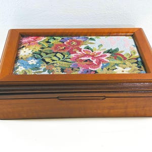 Puede incluir: Una caja rectangular de madera con un acabado marrón pulido y una tapa con bisagras. El centro de la tapa presenta un panel incrustado decorado con un colorido patrón floral de punto de aguja con flores rojas, rosas, amarillas y azules.
