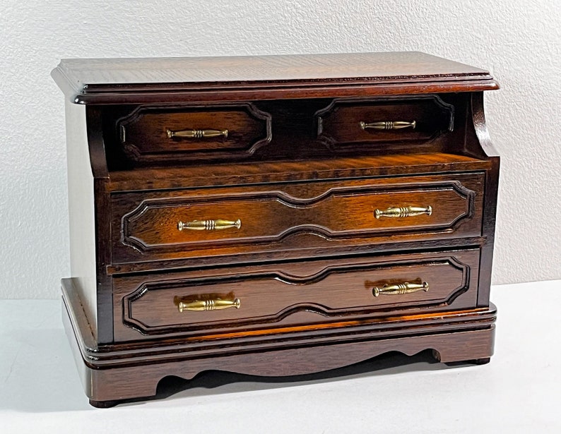 May include: A dark wood jewelry box with three drawers and brass handles. The box has a decorative carved design on the front.