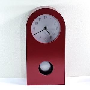 Pode incluir: Um relógio de madeira vermelho com mostrador branco e ponteiros pretos. O relógio tem um pêndulo na parte inferior e tem cerca de 30 cm de altura.