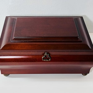 Puede incluir: Una caja rectangular de madera en tono cerezo oscuro con un acabado liso y pulido. La tapa presenta un panel central elevado con bordes biselados. Un pequeño cierre de metal color latón está centrado en la parte frontal, y la caja se apoya sobre cuatro patas.