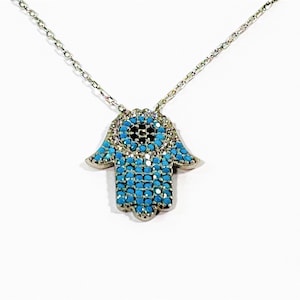 Puede incluir: Un collar de cadena de plata con un colgante de mano de hamsa. El colgante está cubierto de piedras de circonita cúbica azul turquesa y negras.