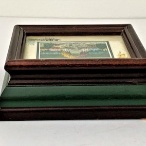 May include: A small framed picture with a green and brown wooden frame and a green base. The picture is of a colorful landscape.