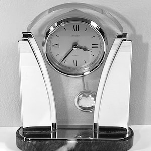 May include: A silver and clear quartz clock with the brand name "Linden" on the clock face. The clock has a pendulum and sits on a black and gray marble base. The clock face has Roman numerals.