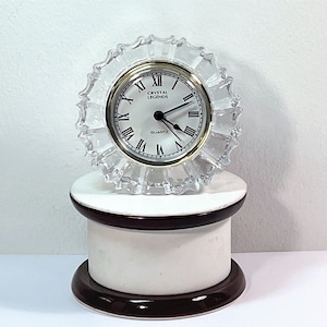 Puede incluir: Un reloj de cuarzo de cristal transparente con borde con volantes, que presenta una esfera blanca con números romanos negros y ribete dorado. El reloj está etiquetado como "Crystal Legends Quartz". Se encuentra sobre un pedestal blanco y marrón.