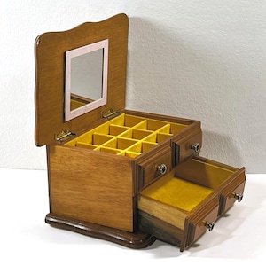Puede incluir: Joyero de madera con tapa con bisagras y un pequeño espejo. La caja tiene múltiples compartimentos y cajones forrados con tela amarilla. El exterior es de color marrón cálido y los herrajes son dorados.