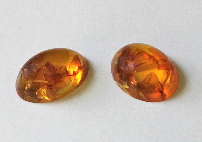 May include: Two oval-shaped amber stones with inclusions of insects. The stones are a light orange color and have a smooth, polished surface.