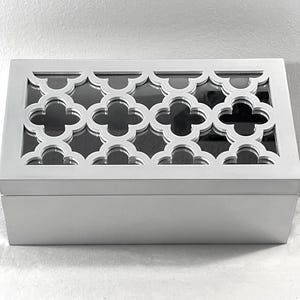 May include: A rectangular, silver-colored box with a decorative, openwork top featuring a repeating quatrefoil pattern. The box has a solid base and a contrasting dark interior, suggesting a storage container.
