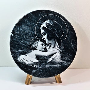 May include: A round, black marble plaque with a white image of the Virgin Mary holding a baby Jesus. The plaque is on a gold stand.