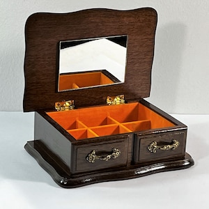 Puede incluir: Una caja de joyería de madera marrón oscuro con una tapa con bisagras y un espejo en el interior. La caja tiene dos cajones con tiradores dorados ornamentados y un interior de terciopelo naranja con compartimentos.