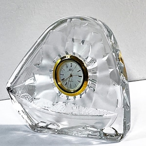 Puede incluir: Un reloj de cristal transparente con una esfera y agujas doradas. El reloj tiene forma de triángulo y presenta un diseño decorativo.