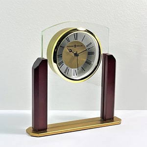 May include: A gold-toned clock with Roman numeral markings on a white face. The clock is encased in a clear glass frame with a gold base and two dark wood supports.