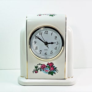 May include: White porcelain clock with a gold trim and a floral design featuring pink roses. The clock face is white with black hands and black numbers.
