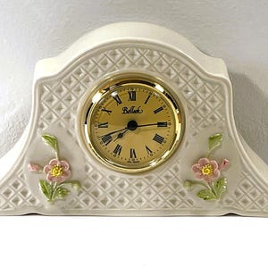 Puede incluir: Reloj de sobremesa de cerámica color crema con esfera dorada y números romanos. El reloj presenta un patrón de diamantes y detalles florales rosas y amarillos. La esfera del reloj tiene la palabra "Belleek".