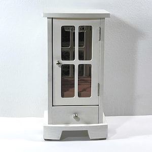 May include: A small, off-white wooden cabinet with a glass-paned door and a drawer. The door has a grid pattern, and a small silver knob is visible. The cabinet sits on a matching base. The interior is visible through the glass.