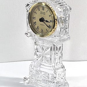 May include: A clear cut crystal mantel clock with a gold-tone bezel and a cream-colored face. The clock features black Roman numerals and the text Crystal Legends and Quartz. The body has a faceted geometric pattern with a rectangular base.