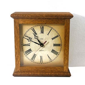 Könnte beinhalten: Eine quadratische Holzuhr mit hellbraunem Rahmen. Das Zifferblatt hat schwarze römische Ziffern und Zeiger mit dem Text "FBN Quartz". Die Uhr ist im Vintage-Stil.