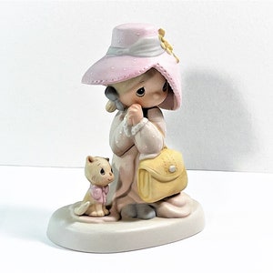 Puede incluir: Una figura de Precious Moments de una niña con sombrero y vestido rosa, sosteniendo un bolso amarillo. Un pequeño gatito con un lazo rosa está sentado a su lado. La figura está sobre una base blanca.
