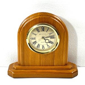 Op de afbeelding: Een houten quartz klok met een goudkleurige wijzerplaat en Romeinse cijfers. De klok heeft een lichtbruin houten frame met een gewelfde bovenkant en een rechthoekige basis. De wijzerplaat leest "Reflections Quartz".