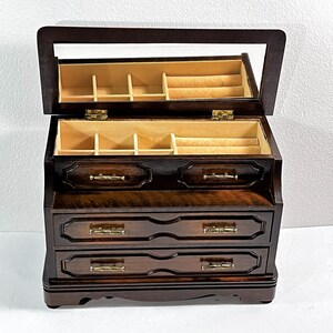 May include: A dark wood jewelry box with a hinged lid and a mirror inside. The box has three drawers with ornate carvings and brass handles.