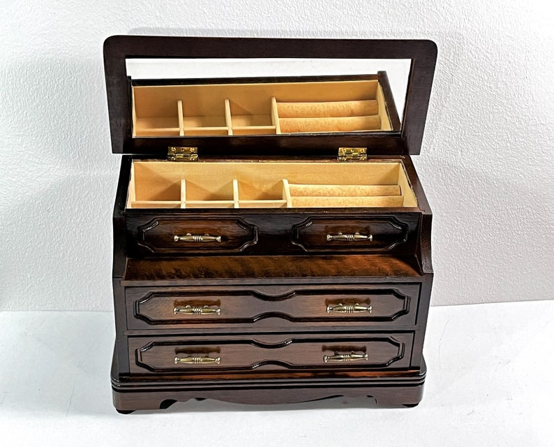 May include: A dark wood jewelry box with a hinged lid and a mirror inside. The box has three drawers with ornate carvings and brass handles.