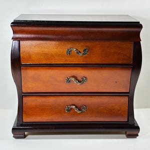 Puede incluir: Una pequeña cómoda de madera con tres cajones, que presenta un marco marrón oscuro y frentes de cajón de color marrón claro. Cada cajón está equipado con un tirador de metal curvado y ornamentado de color latón con acabado pulido.