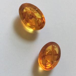 May include: Two oval-shaped amber buttons with a translucent, honey-colored appearance. The buttons have a textured surface with a pattern of inclusions.