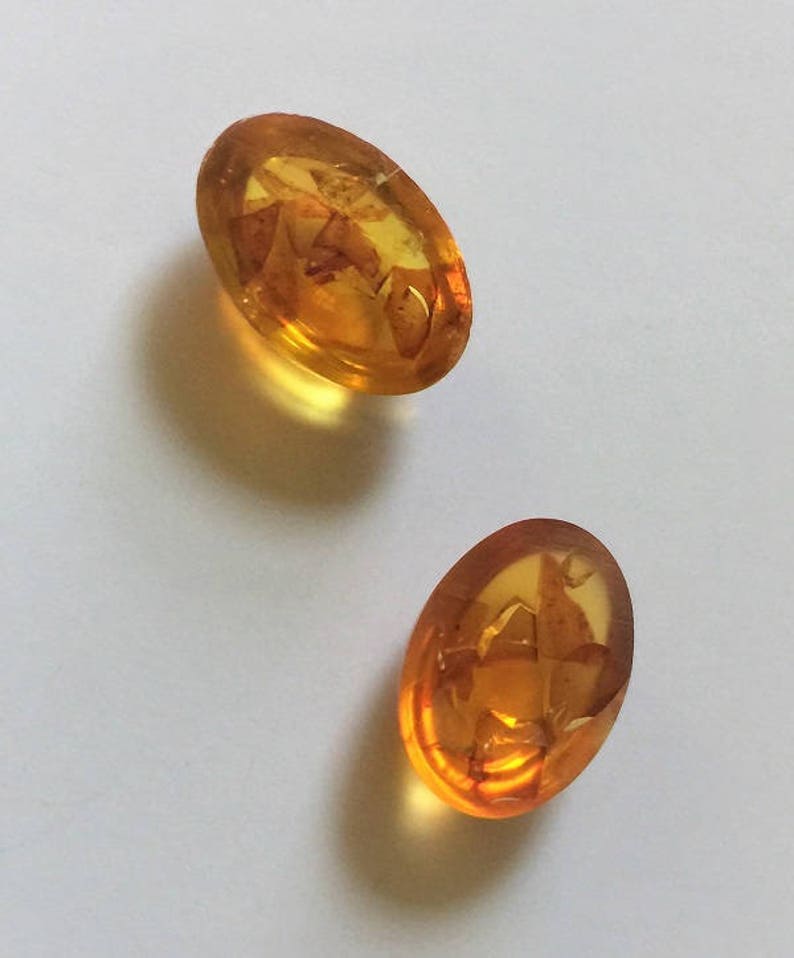 May include: Two oval-shaped amber buttons with a translucent, honey-colored appearance. The buttons have a textured surface with a pattern of inclusions.