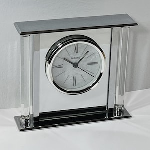 Puede incluir: Un reloj de escritorio Bulova con marco plateado y parte trasera espejada. La esfera es blanca con números romanos y manecillas negras. El reloj es rectangular con soportes verticales transparentes.